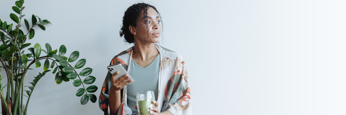 Woman sitting next to plant, holding phone in one hand and green juice in the other and looking to the side